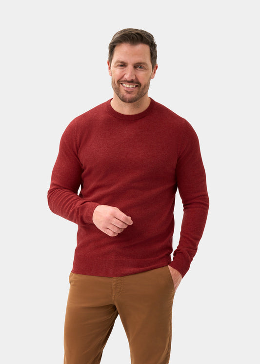 Man wears Brisbane Geelong Wool Crew Neck Jumper in Poppy Melange and brown pants, smiling with one hand in his pocket.