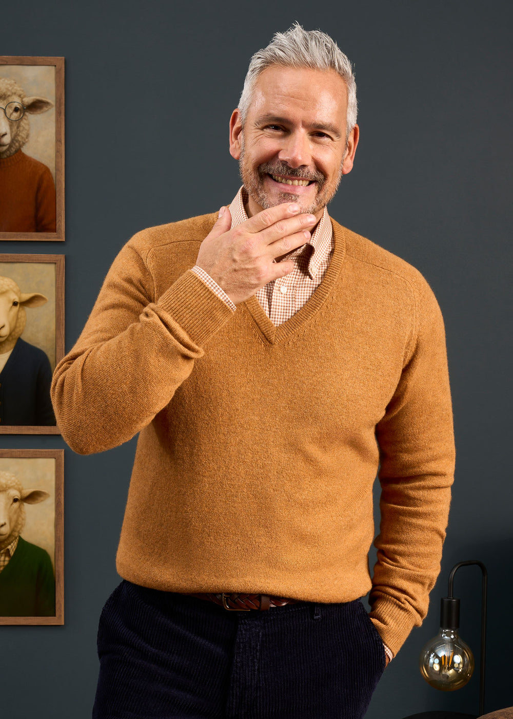 Man with gray hair wears Hampshire Lambswool Vee Neck Jumper in Gazelle, standing before sheep portraits on dark wall.