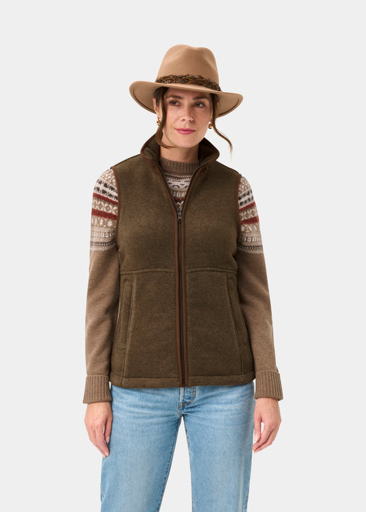 Woman in a tan hat, patterned sweater, Aylsham Ladies Fleece Gilet in Pecan, and jeans stands against a white background.