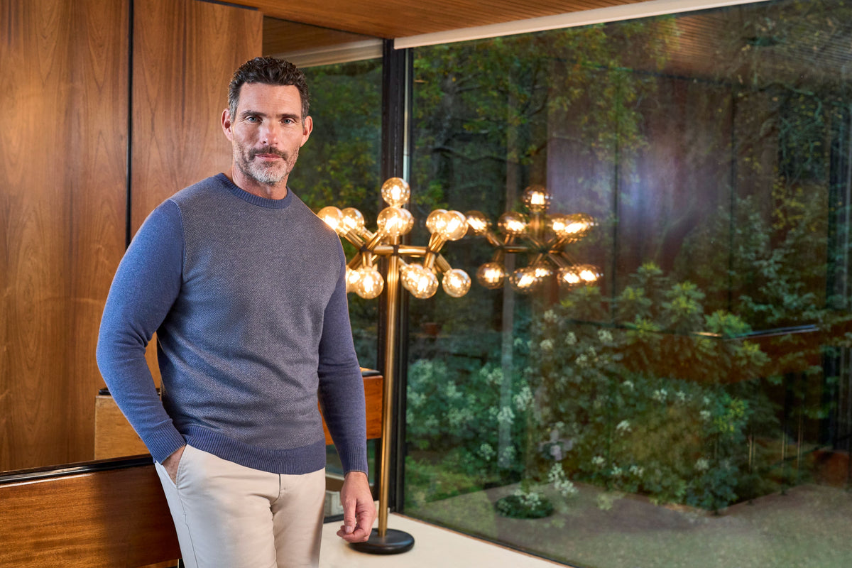 A man wearing a slate blue crewneck sweater and beige trousers stands indoors next to a modern multi-arm chandelier with glowing bulbs, with a large window showing a garden view behind him.