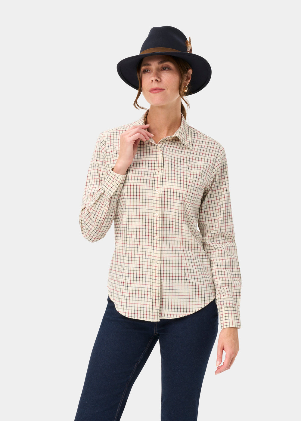 Woman in Bromford Ladies Cotton Shirt In Red Check poses with a dark hat, one hand near collar, on white background.