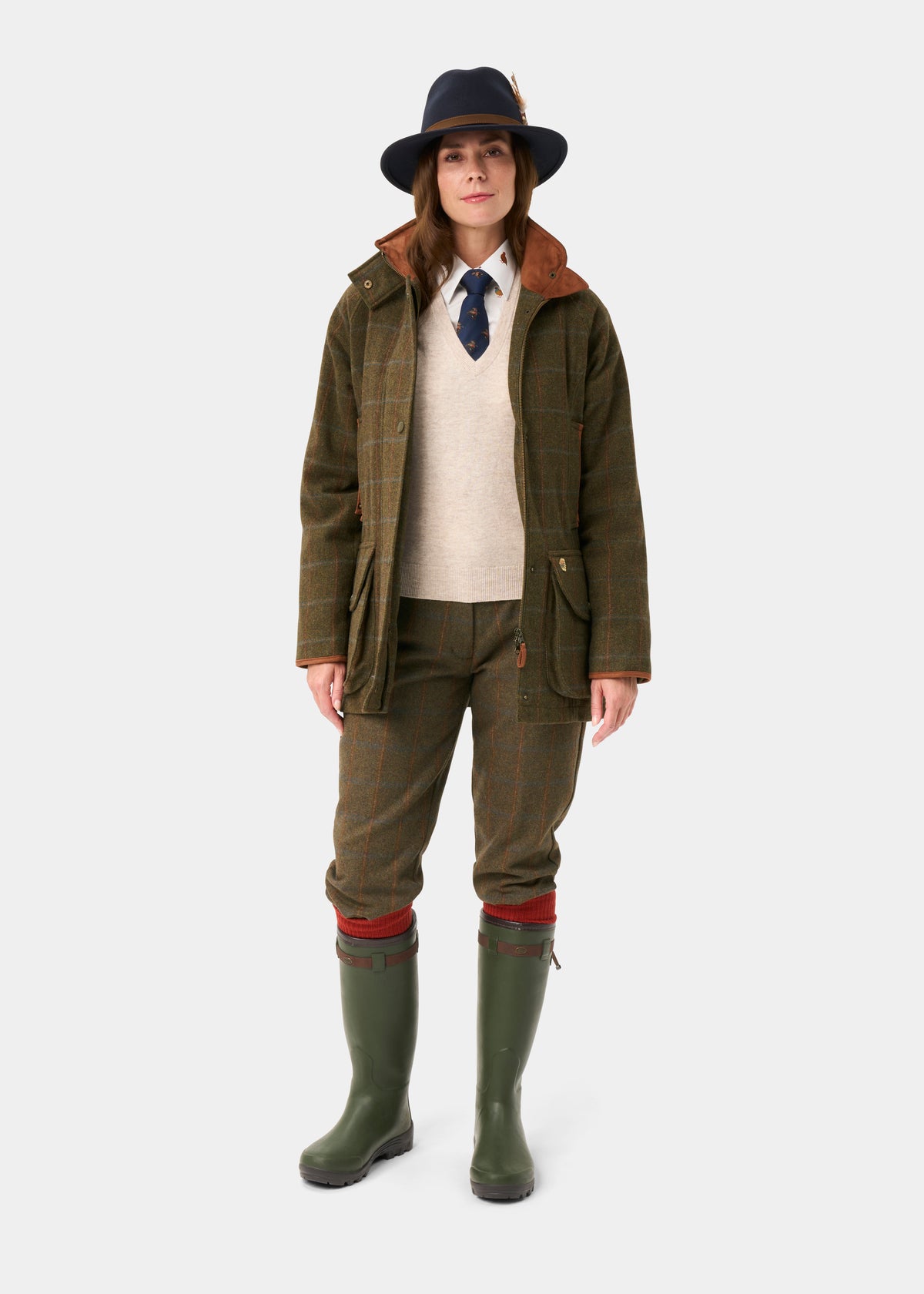 Woman models Combrook Ladies Tweed Waterproof Shooting Coat in Willow with hat and green boots on white background.