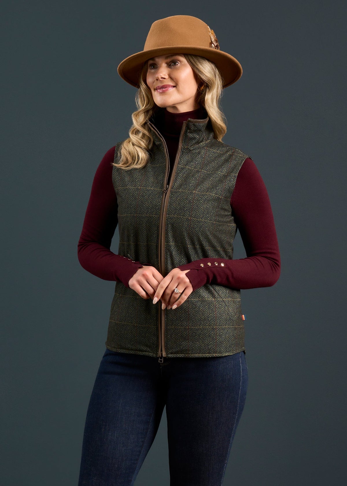 Woman in a brown hat, burgundy top, and Myrtle Didsmere Ladies Technical Tweed Gilet poses against a dark background.