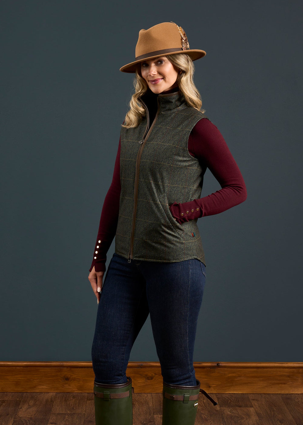 Woman in maroon top, Didsmere Ladies Technical Tweed Effect Gilet in Myrtle, jeans, and boots poses on wood floor.