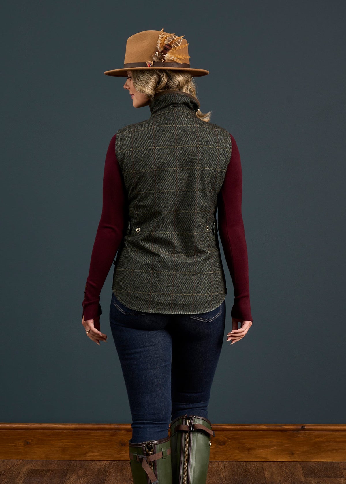 Woman facing away, wearing brown feathered hat, maroon sleeves, jeans, green boots & Didsmere Myrtle Gilet.