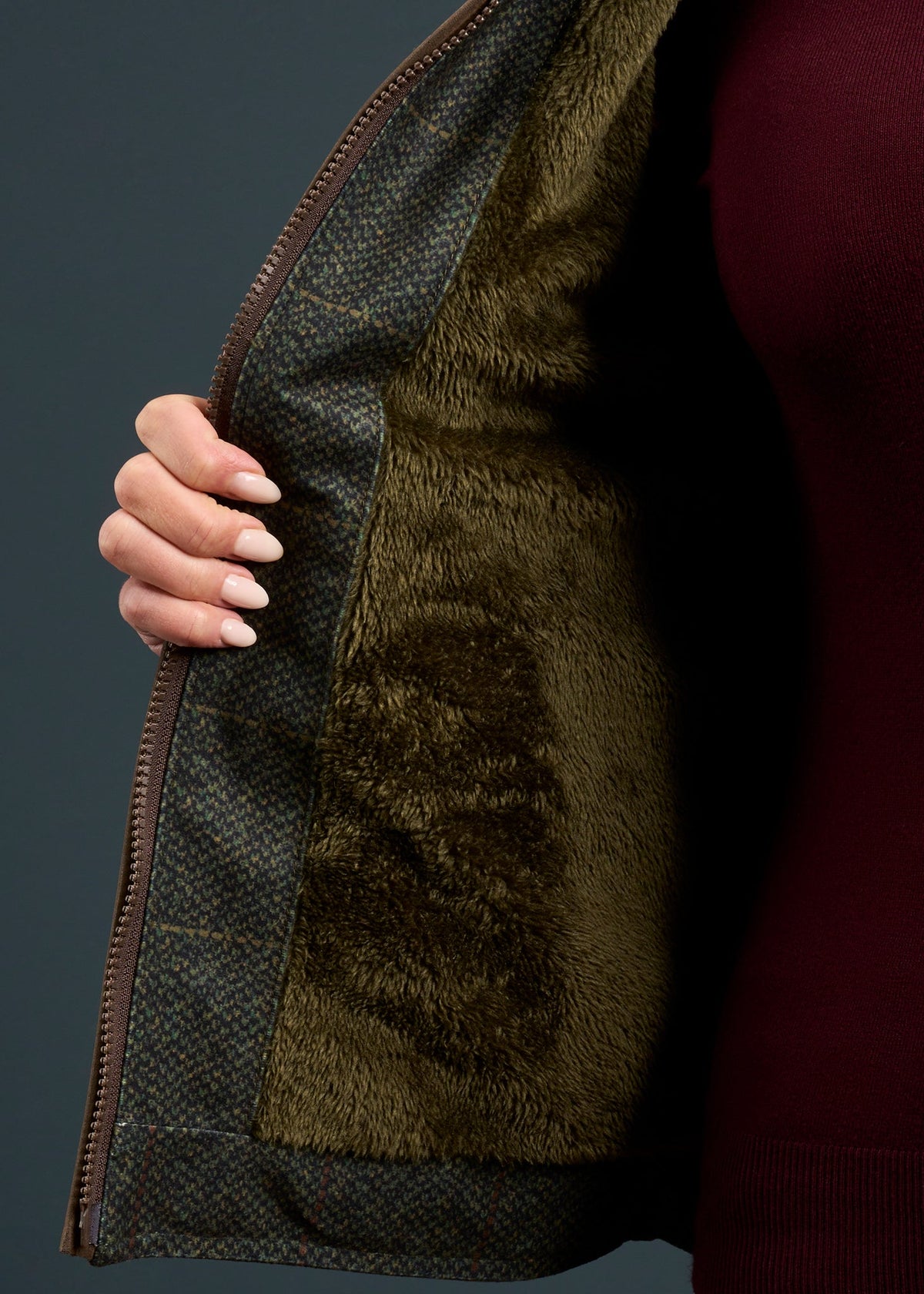 A woman opens a Didsmere Technical Tweed Effect Gilet in Myrtle, showing its faux fur lining over a burgundy top.