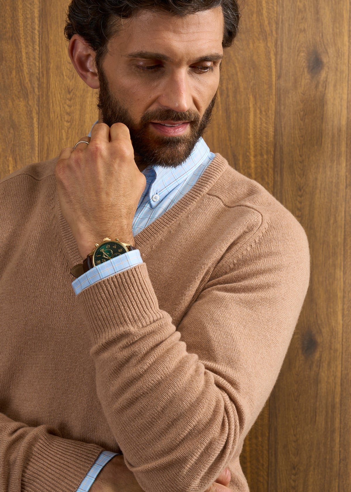 Man wears Hampshire Lambswool Vee Neck Jumper in Camel over blue shirt, gold watch visible, wood panel backdrop.