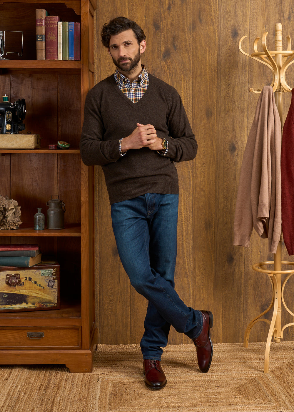 Man in Hampshire Lambswool Vee Neck Jumper In Cocoa leans against bookcase in a cozy, wood-paneled room.
