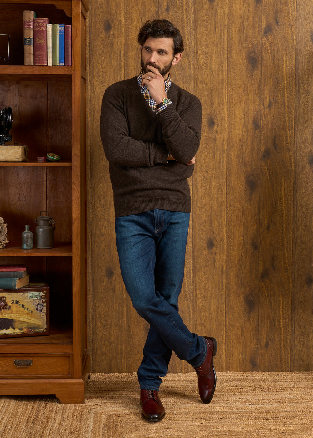 Man in a Hampshire Lambswool Vee Neck Jumper in Cocoa stands by a bookshelf, looking thoughtful with hand on chin.