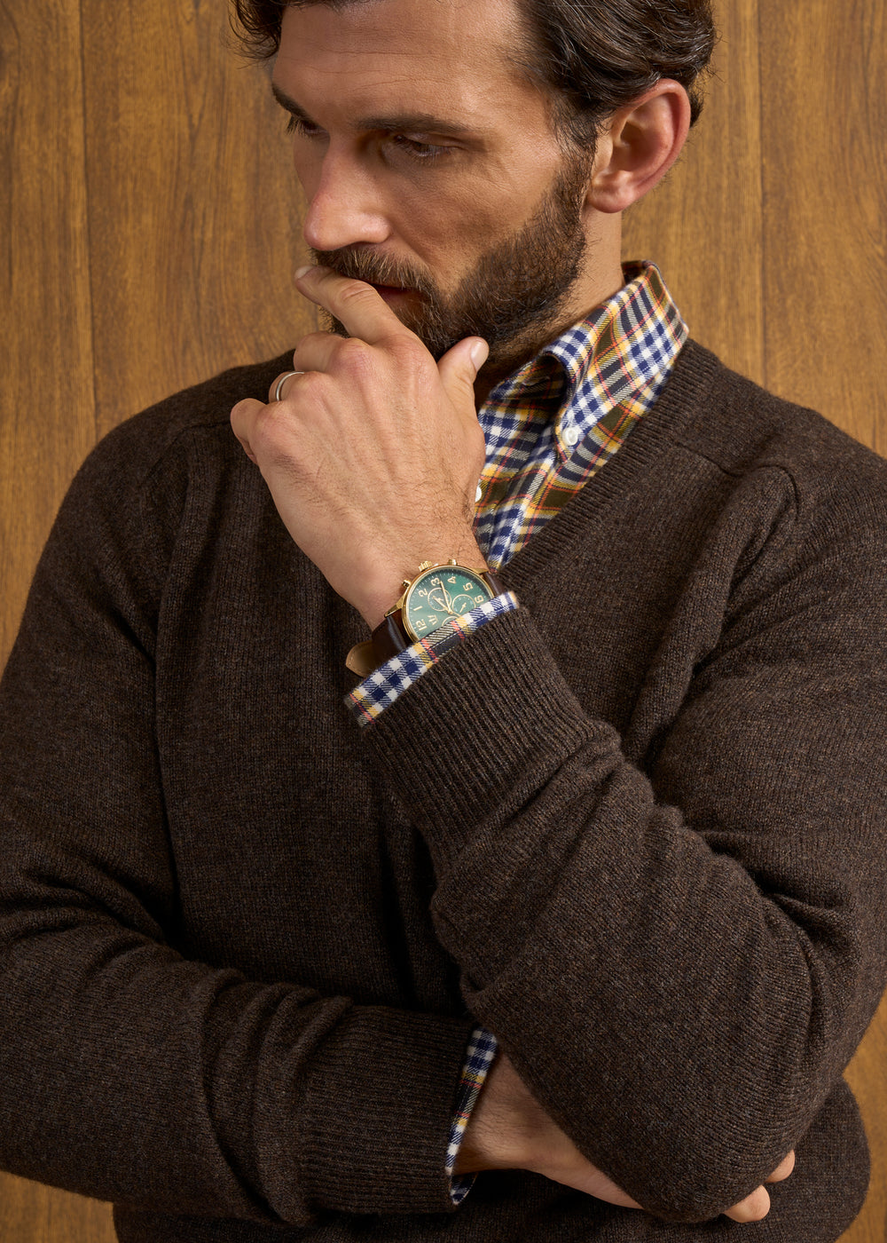 Man in Hampshire Lambswool Vee Neck Jumper, plaid shirt & watch stands by a wood-paneled wall.