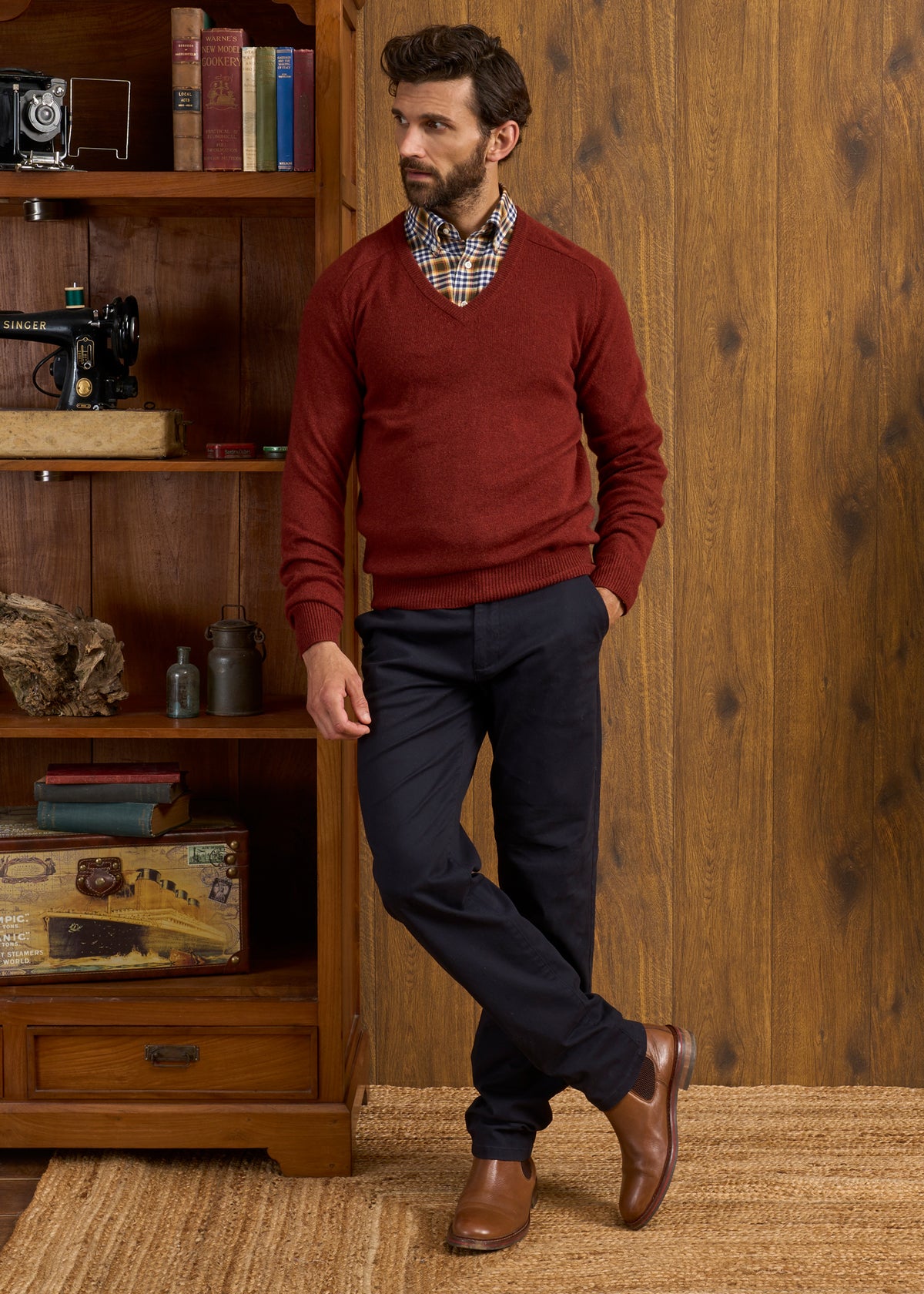 Man in a Hampshire Lambswool Vee Neck Jumper in Sienna, plaid shirt, and navy pants stands by a wooden bookshelf.