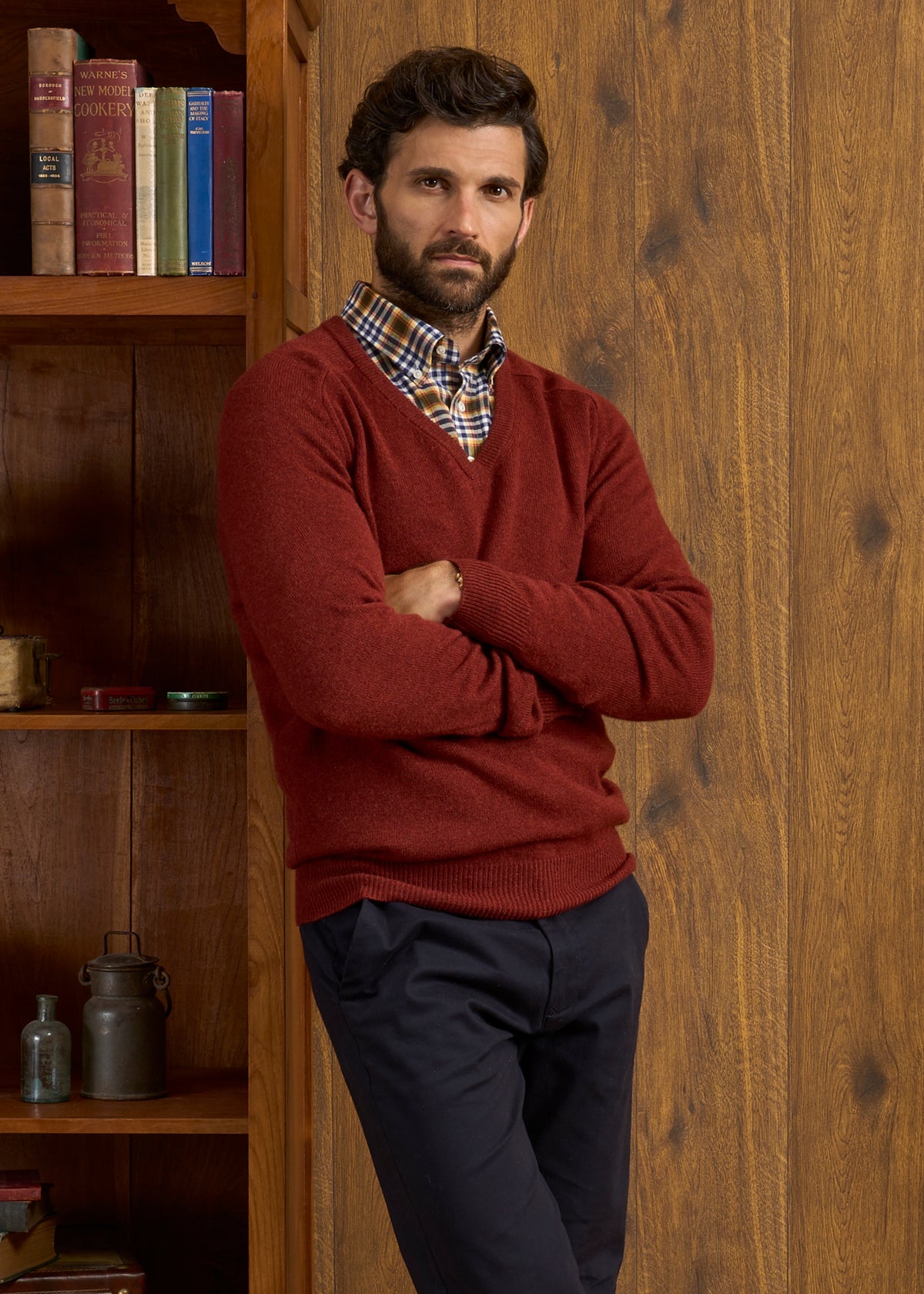 Man in sienna Hampshire Lambswool Vee Neck Jumper stands by bookshelf, red sweater and plaid shirt layered.