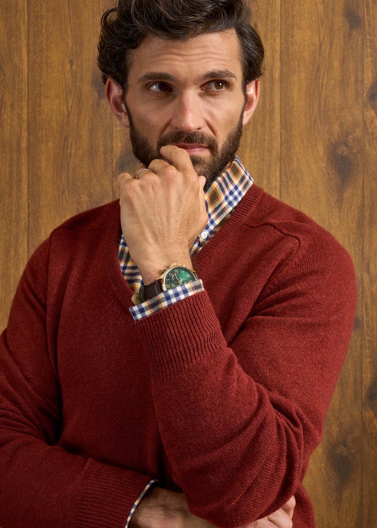Man in Hampshire Lambswool Vee Neck Jumper in Sienna stands thoughtfully, hand on chin, by a wooden background.
