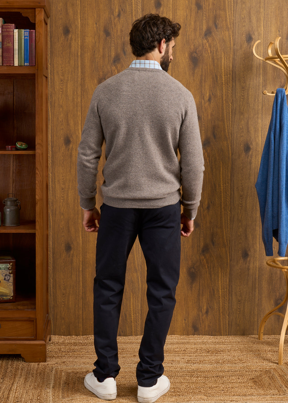 Man indoors, facing away, in Hampshire Lambswool Vee Neck Jumper in Vole; navy pants, white shoes; wood panel background.