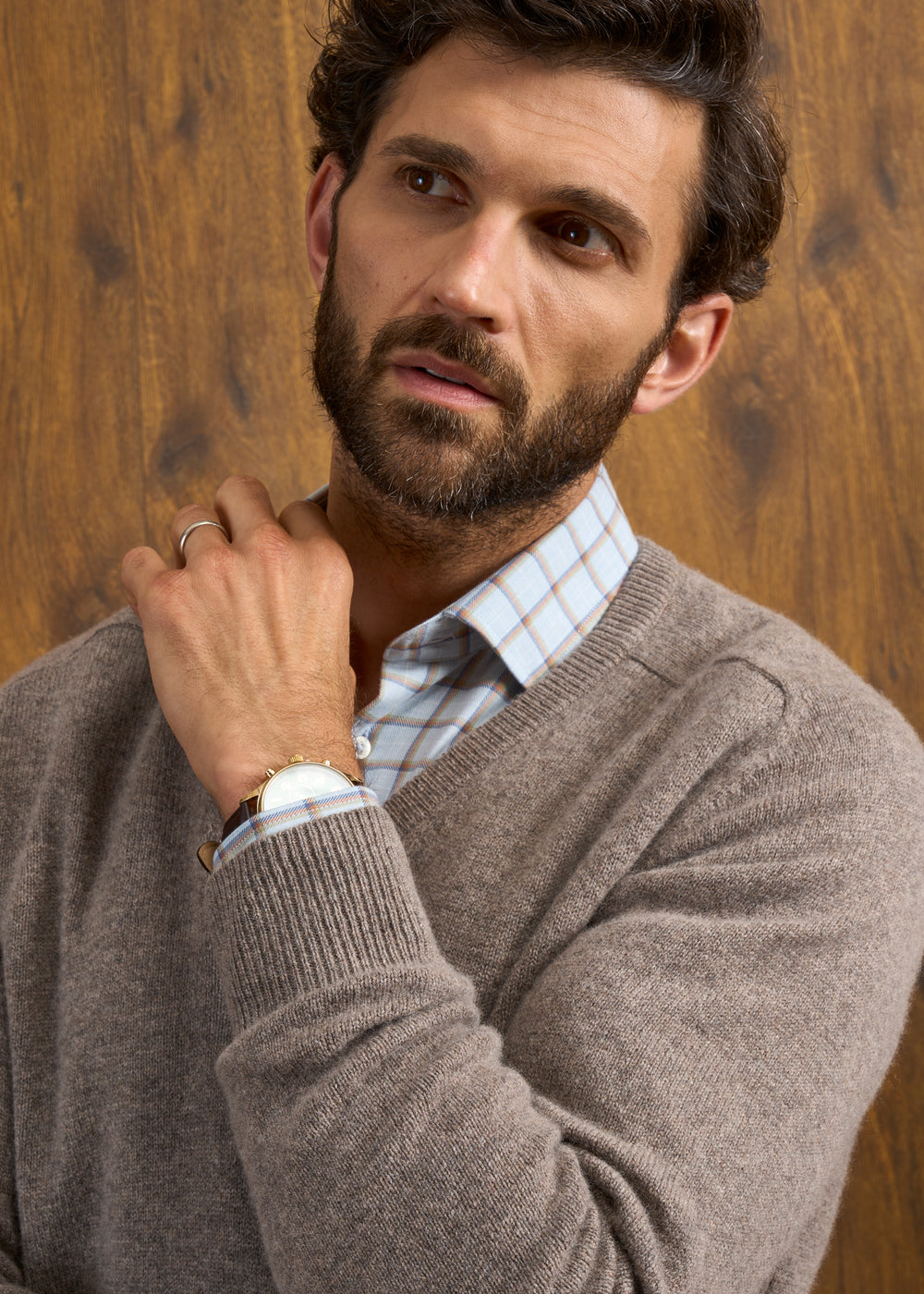 Man with brown hair and beard wears Hampshire Lambswool Vee Neck Jumper in Vole, standing by a wooden wall.