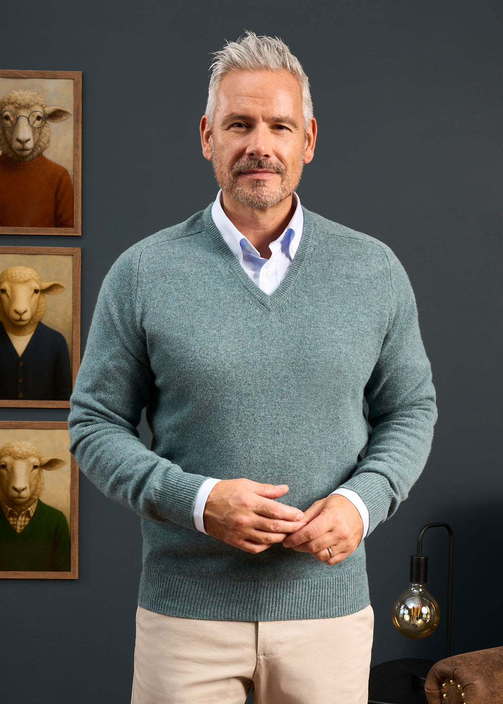 A man with gray hair wears a Hampshire Lambswool Vee Neck Jumper in Caspian; sheep art and a lamp are behind him.