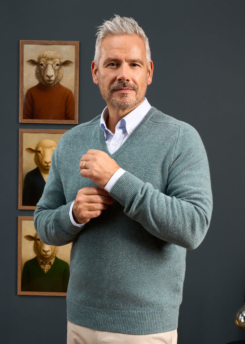 A man in a Hampshire Lambswool Vee Neck Jumper in Caspian adjusts his sleeve near three framed sheep portraits.
