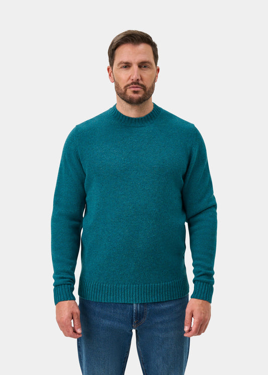 Man wears Kinnadie Supersoft Shetland Wool Crew Neck Jumper in Larkspur and blue jeans, against white background.