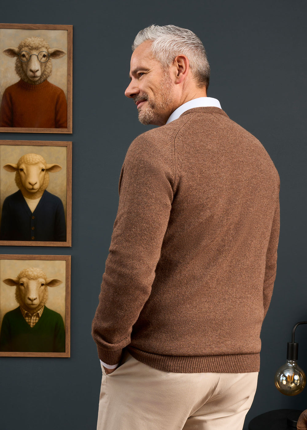 A man in a Hampshire Lambswool Vee Neck Jumper in Tobacco stands by three sheep portraits on a dark wall.