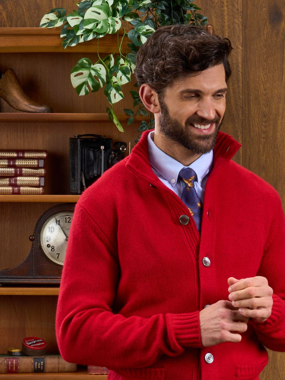 Smiling man in a Landford Lambswool Jumper stands before shelves with books, a clock, and a plant.