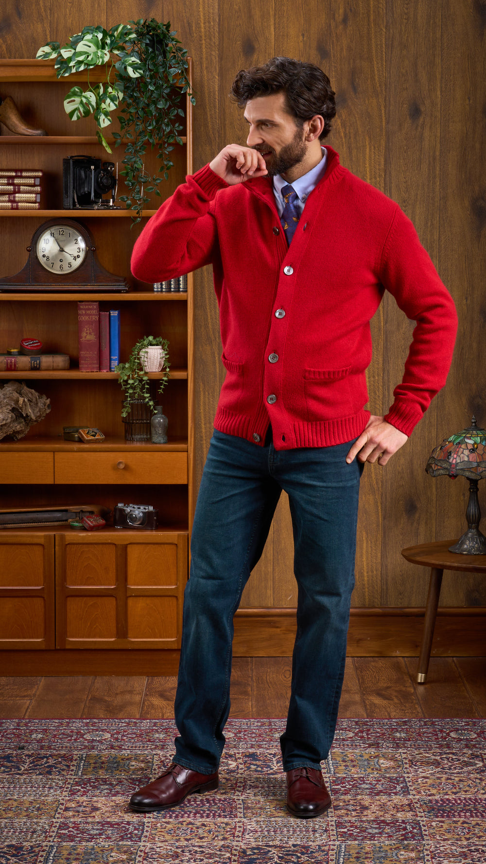 A bearded man in a red Landford lambswool jumper, blue shirt, tie, and jeans stands in a vintage study.