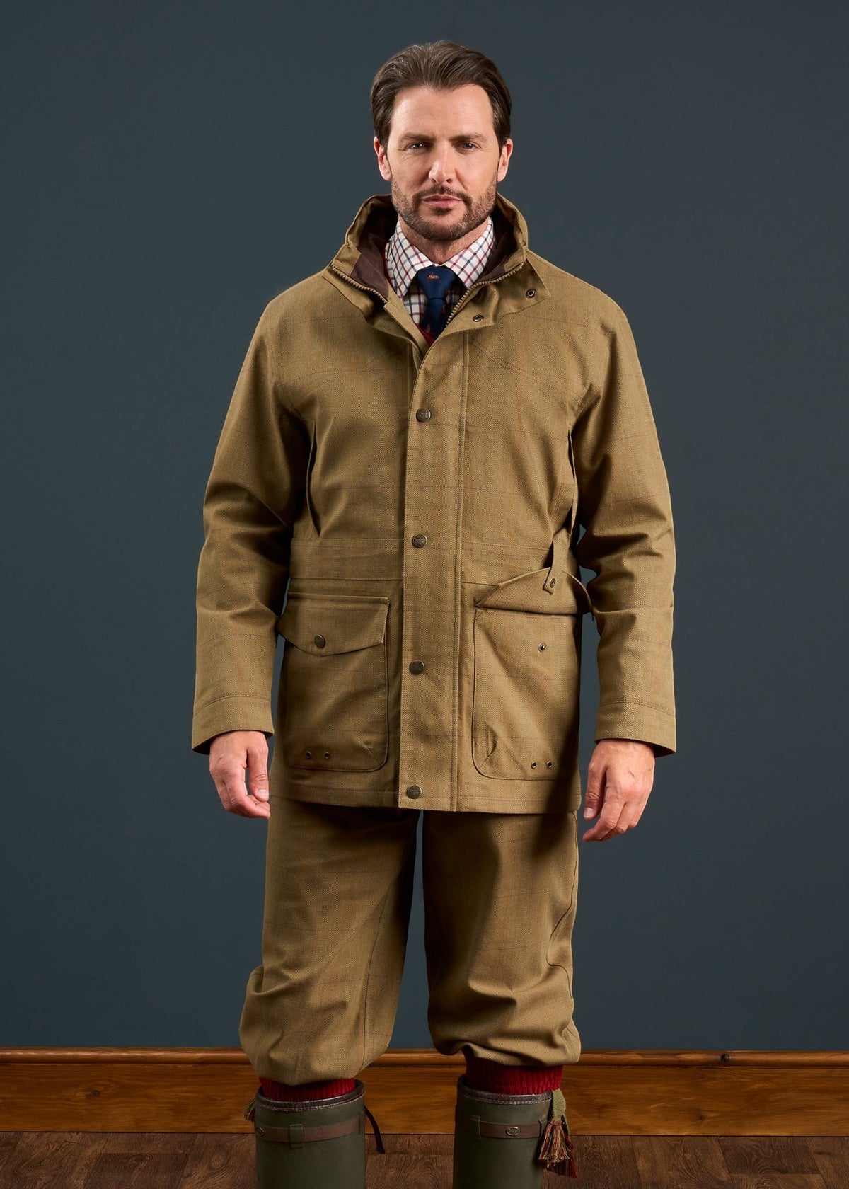 Man in Axford Men’s Waterproof Lightweight Field Coat in Aspen, checked shirt, tie, and boots stands against dark backdrop.