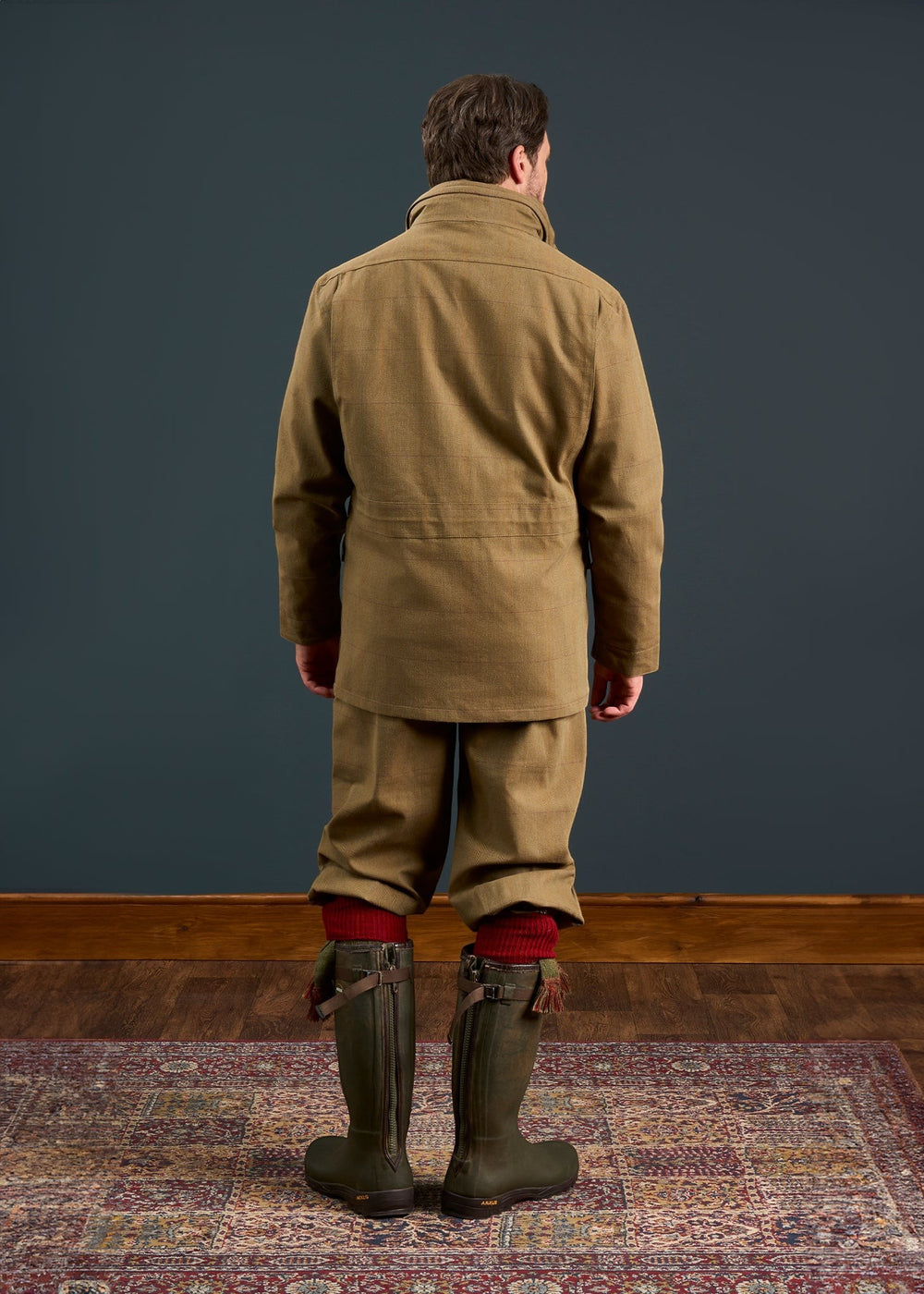 A man in vintage boots stands on a rug, modeling the Axford Men’s Waterproof Field Coat in Aspen, against a dark wall.
