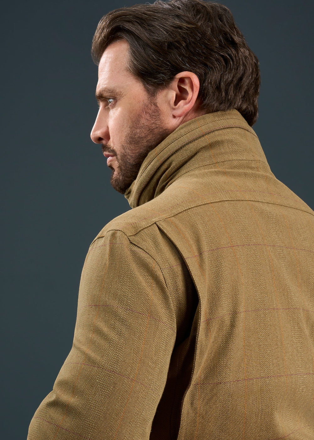 Man with brown hair and beard wears Axford Men’s Waterproof Field Coat in Aspen (Shooting Fit), facing left on gray.