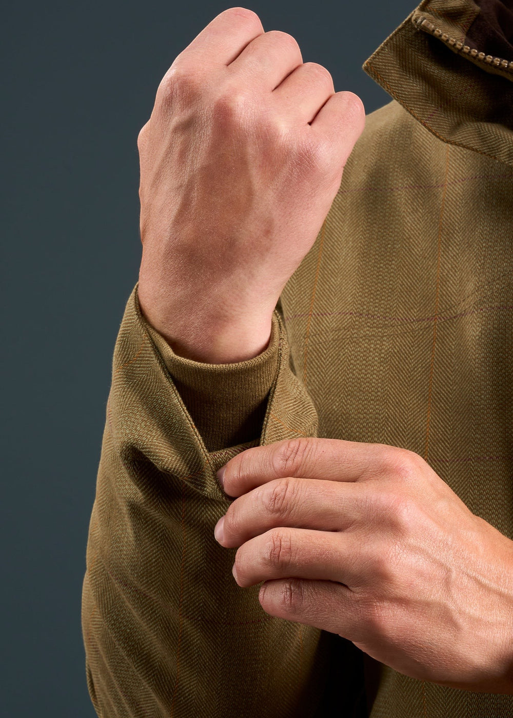 A man adjusts the cuff of an Axford Men’s Aspen Waterproof Field Coat (Shooting Fit) against a dark background.