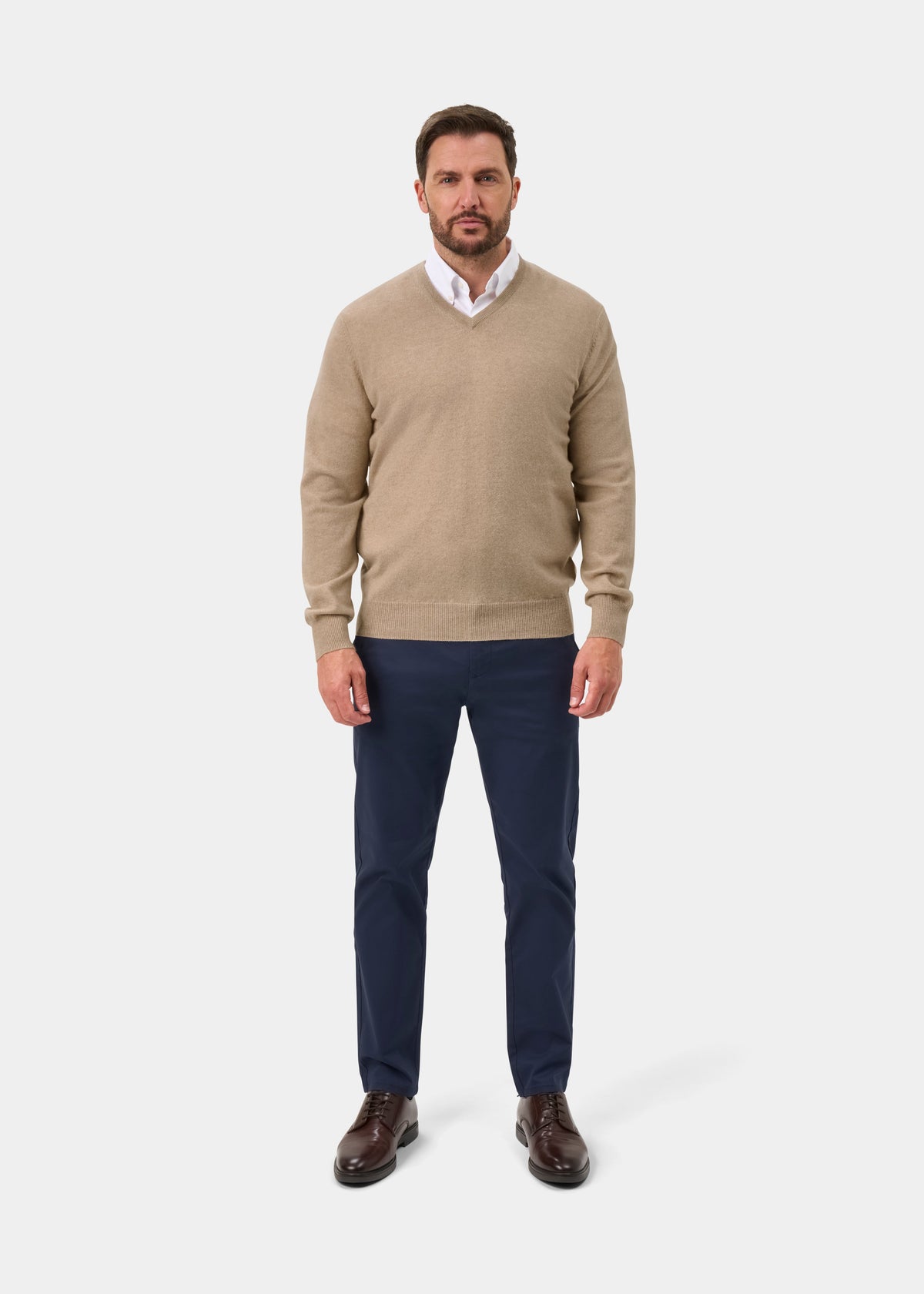 Man wears Haddington Pure Cashmere V-Neck Jumper in Linen over a white shirt, navy pants, and brown shoes.