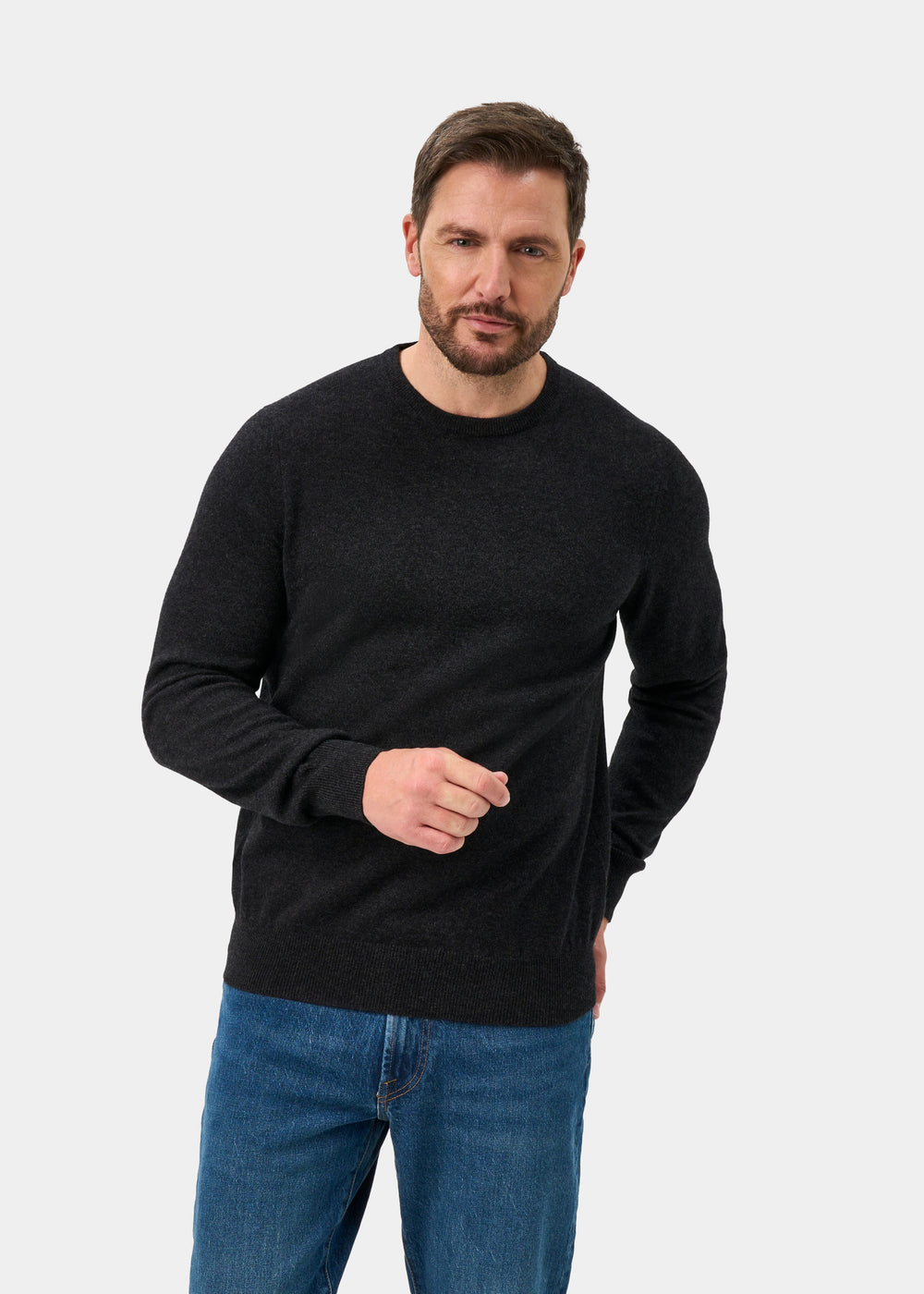Man in a charcoal Brisbane Geelong Wool Crew Neck Jumper and blue jeans stands against a light background, looking ahead.