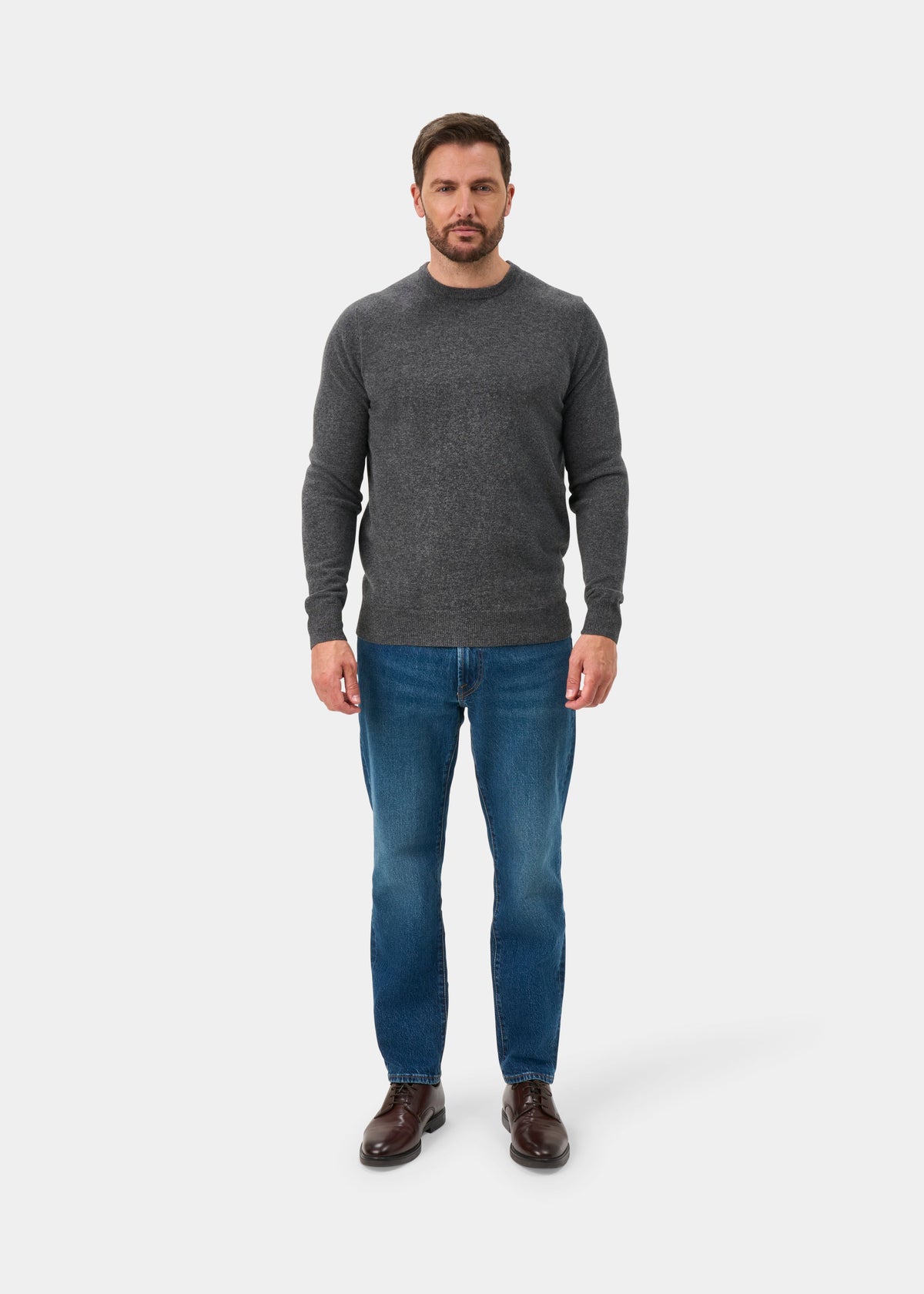 Man wears Brisbane Geelong Wool Crew Neck Jumper in Derby, blue jeans, brown shoes, against a plain white background.