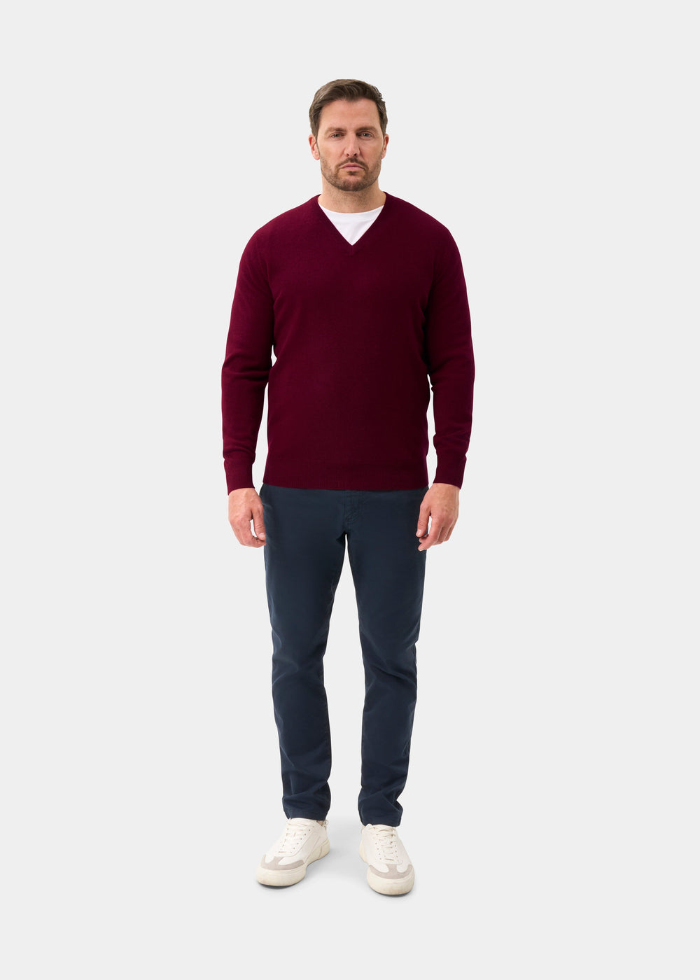 Man in Albury Geelong Wool V-Neck Jumper in Claret, white shirt, navy pants, and white sneakers on light background.