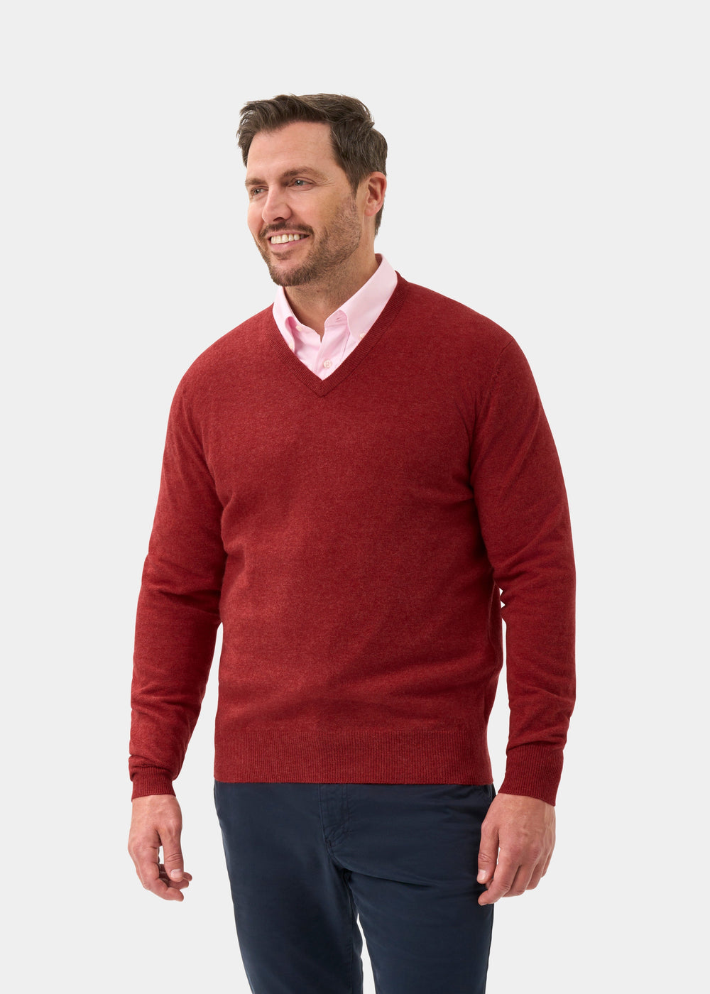 Smiling man in Albury Geelong Wool V-Neck Jumper in Poppy Melange stands against a plain light gray background.