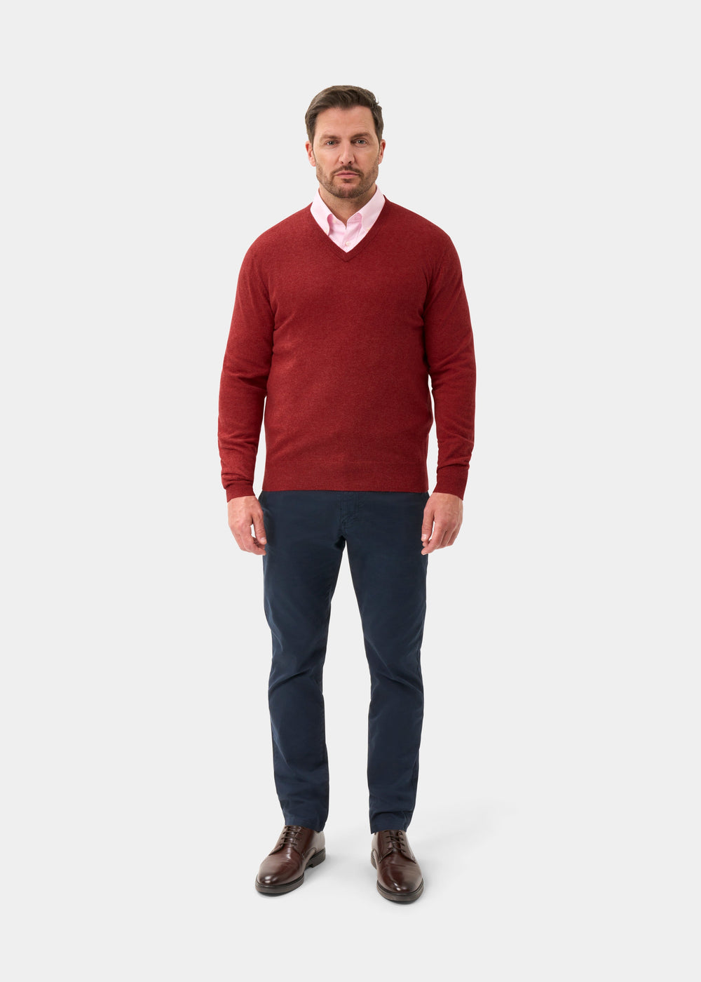 Man wears Albury Geelong Wool V-Neck Jumper in Poppy Melange over pink shirt, navy pants, and brown shoes.