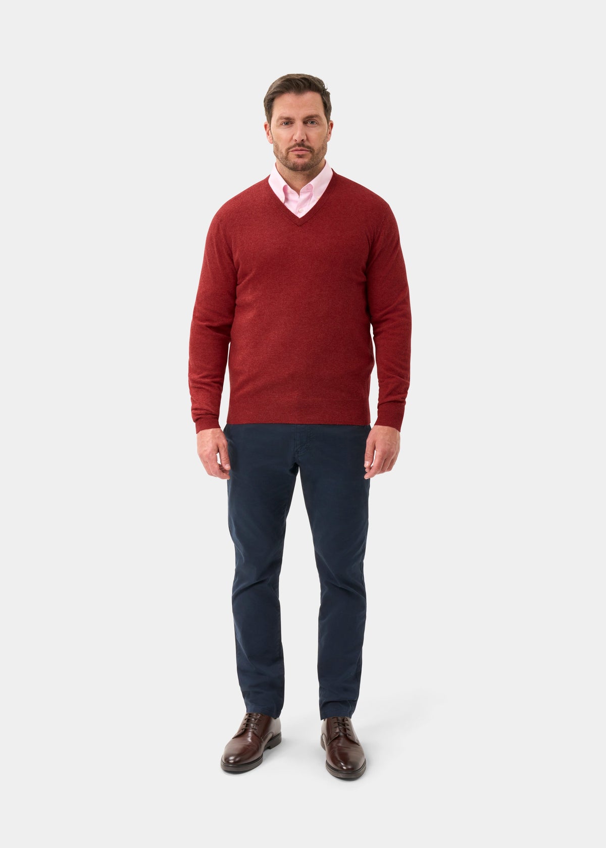 Man wears Albury Geelong Wool V-Neck Jumper in Poppy Melange over pink shirt, navy pants, and brown shoes.