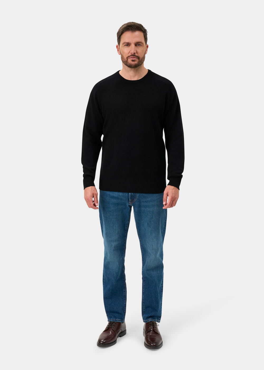 Man facing forward in Dorset Lambswool Crew Neck Jumper in Black, blue jeans, and brown shoes on a plain background.