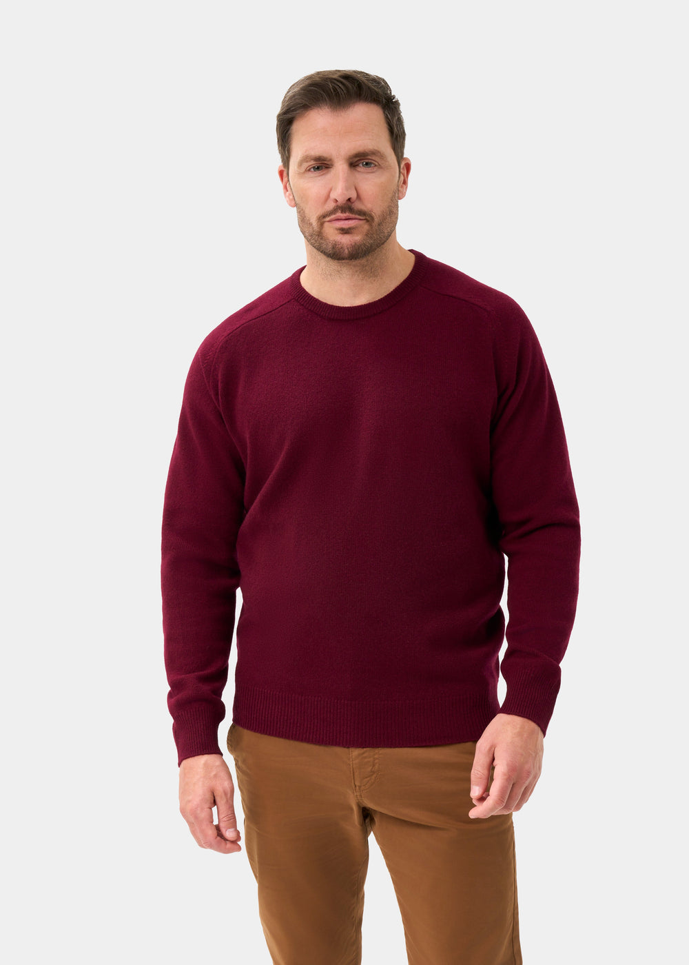 Man wears Dorset Lambswool Crew Neck Jumper in Bordeaux, classic fit, with brown pants against a light gray background.