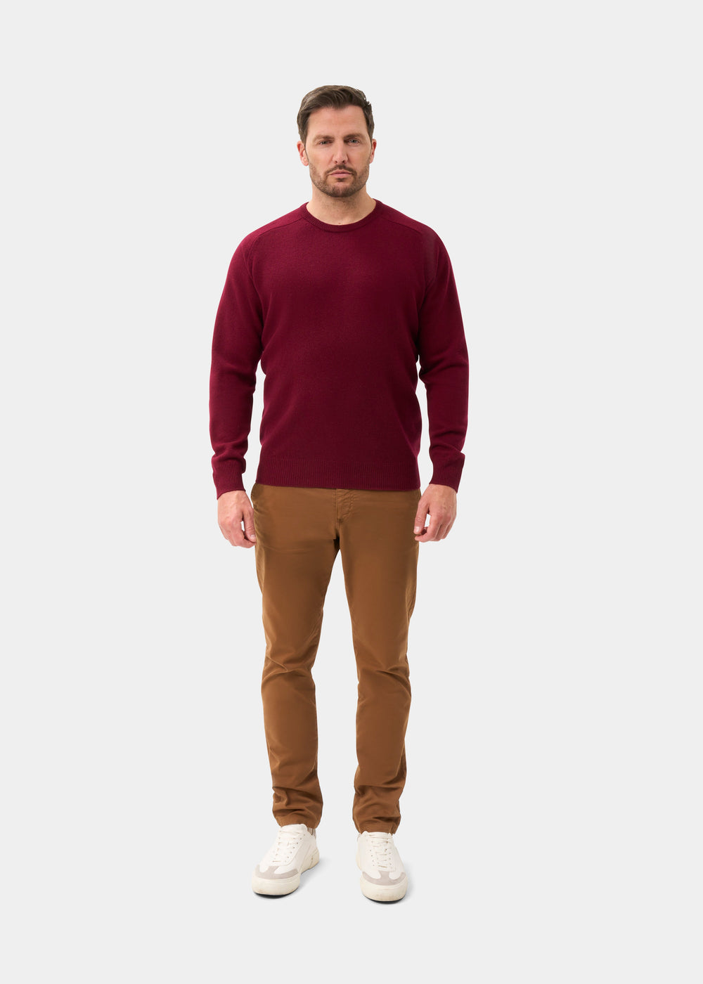 Man in Dorset Lambswool Crew Neck Jumper in Bordeaux, tan pants, and white sneakers stands by a light background.