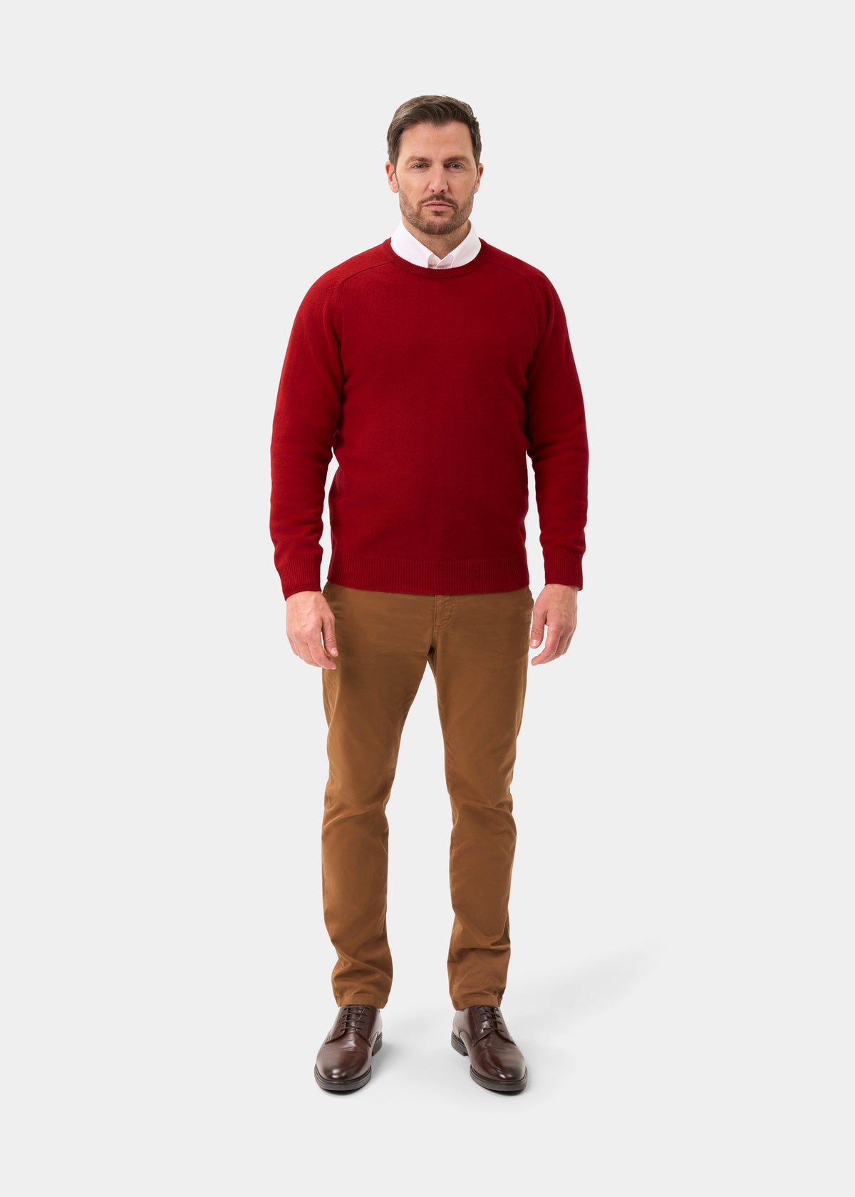Man in Dorset Lambswool Crew Neck Jumper in Dubonnet, white shirt, brown pants and shoes on a plain light background.