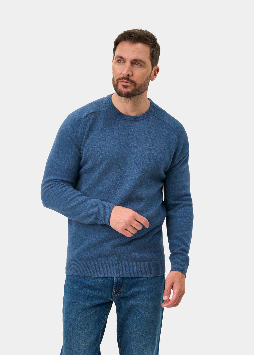 Man wears Lenzie Lambswool Crew Neck Jumper in Jeans, regular fit, against a plain background, looking to the side.