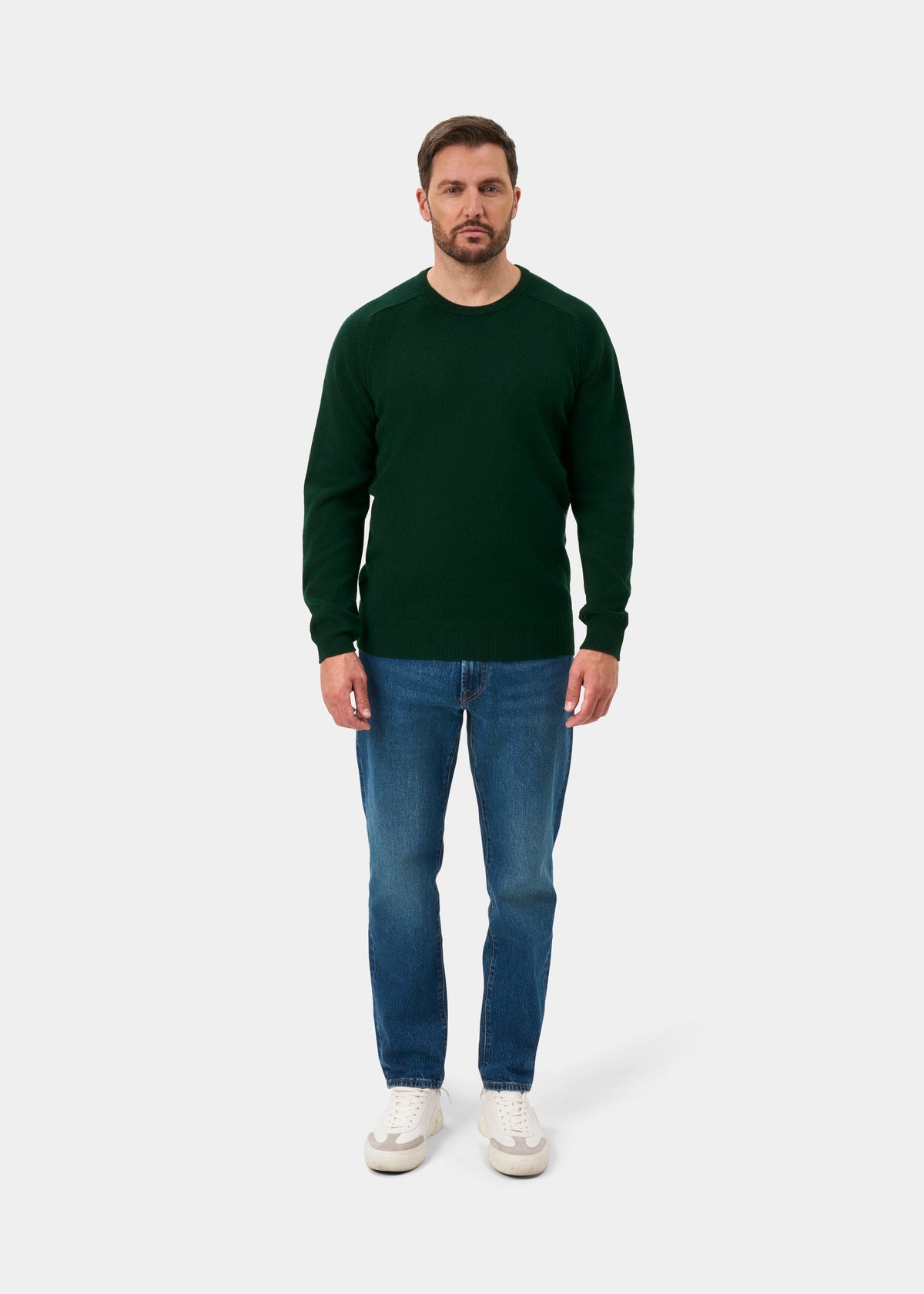 Man in a Dorset Lambswool Crew Neck Jumper in Tartan Green, blue jeans, and white sneakers on a white background.
