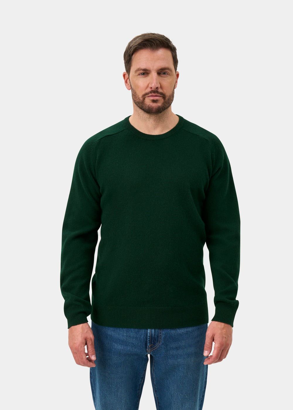 Man wears Dorset Lambswool Crew Neck Jumper in Tartan Green, classic fit, with blue jeans against a plain background.