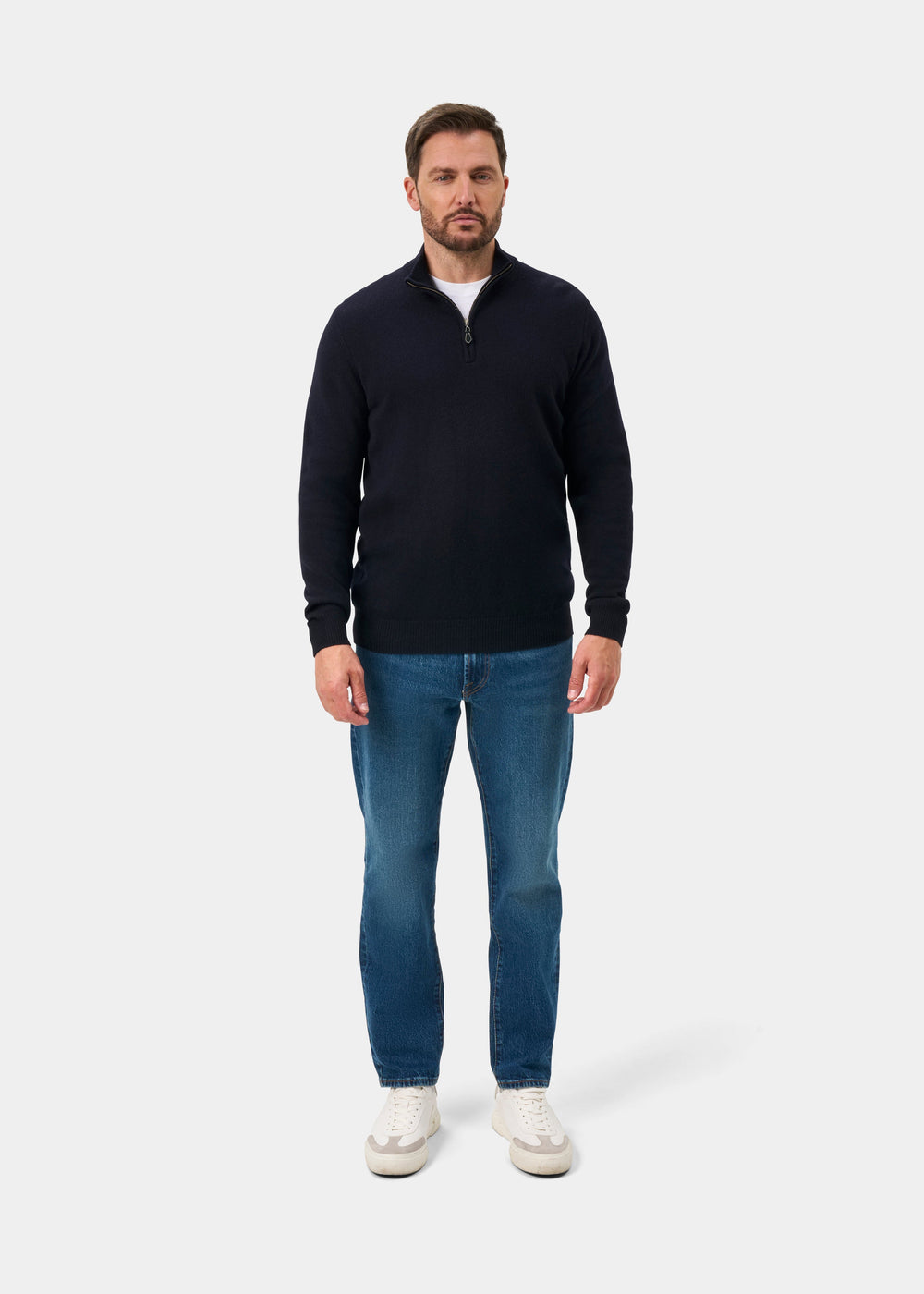 A man wearing a Sutherland Lambswool Quarter Zip Jumper In Navy over a white t-shirt, blue jeans, and white sneakers.