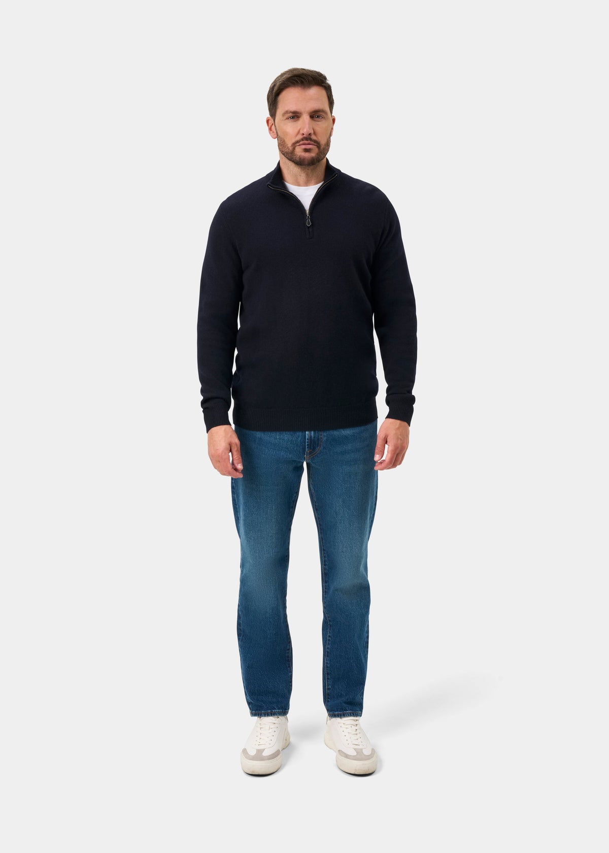 A man wearing a Sutherland Lambswool Quarter Zip Jumper In Navy over a white t-shirt, blue jeans, and white sneakers.