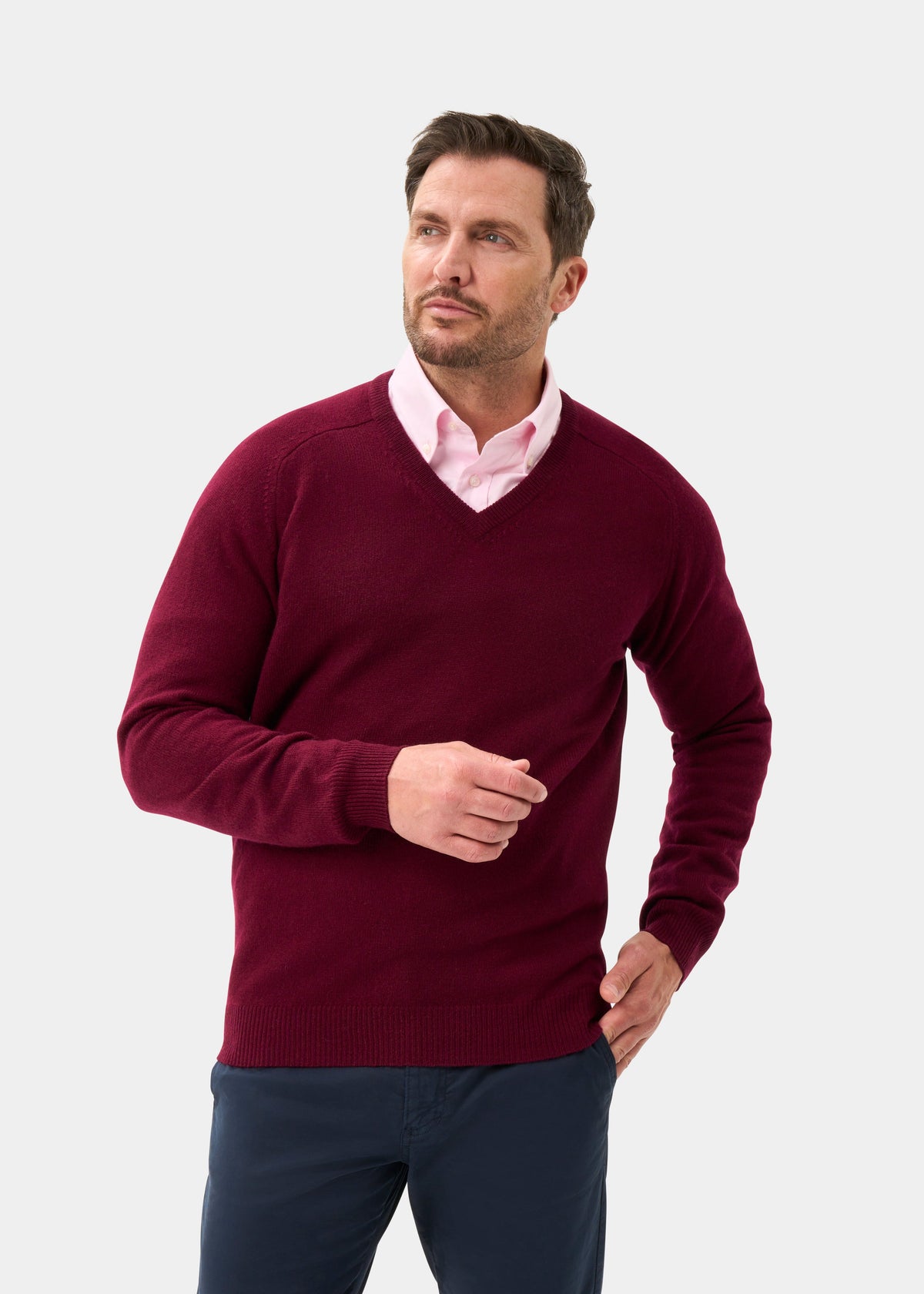 Man wears Kilsyth Lambswool V-Neck Jumper in Bordeaux over pink shirt and dark pants against plain background.