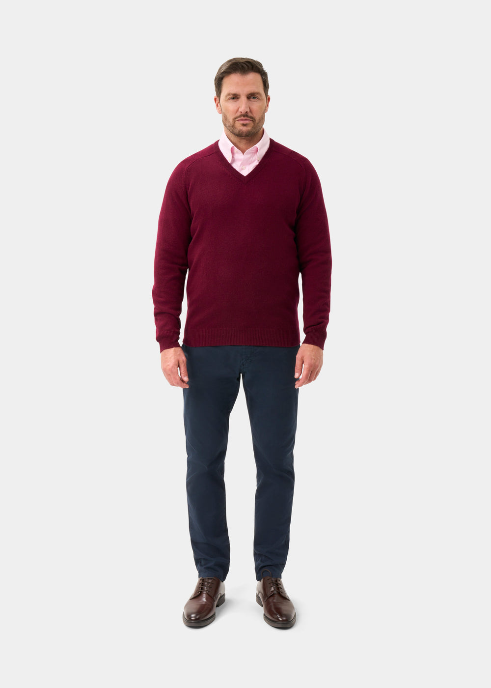 Man in Kilsyth Lambswool V-Neck Jumper in Bordeaux, pink shirt, navy pants, and brown shoes on white background.
