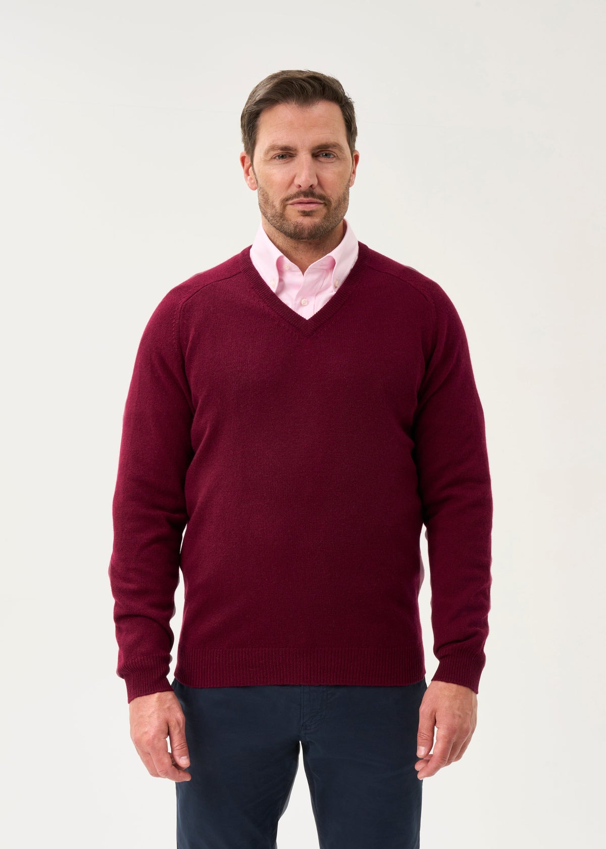 Man wears Kilsyth Lambswool V-Neck Jumper in Bordeaux over pink shirt and dark pants, with light background.