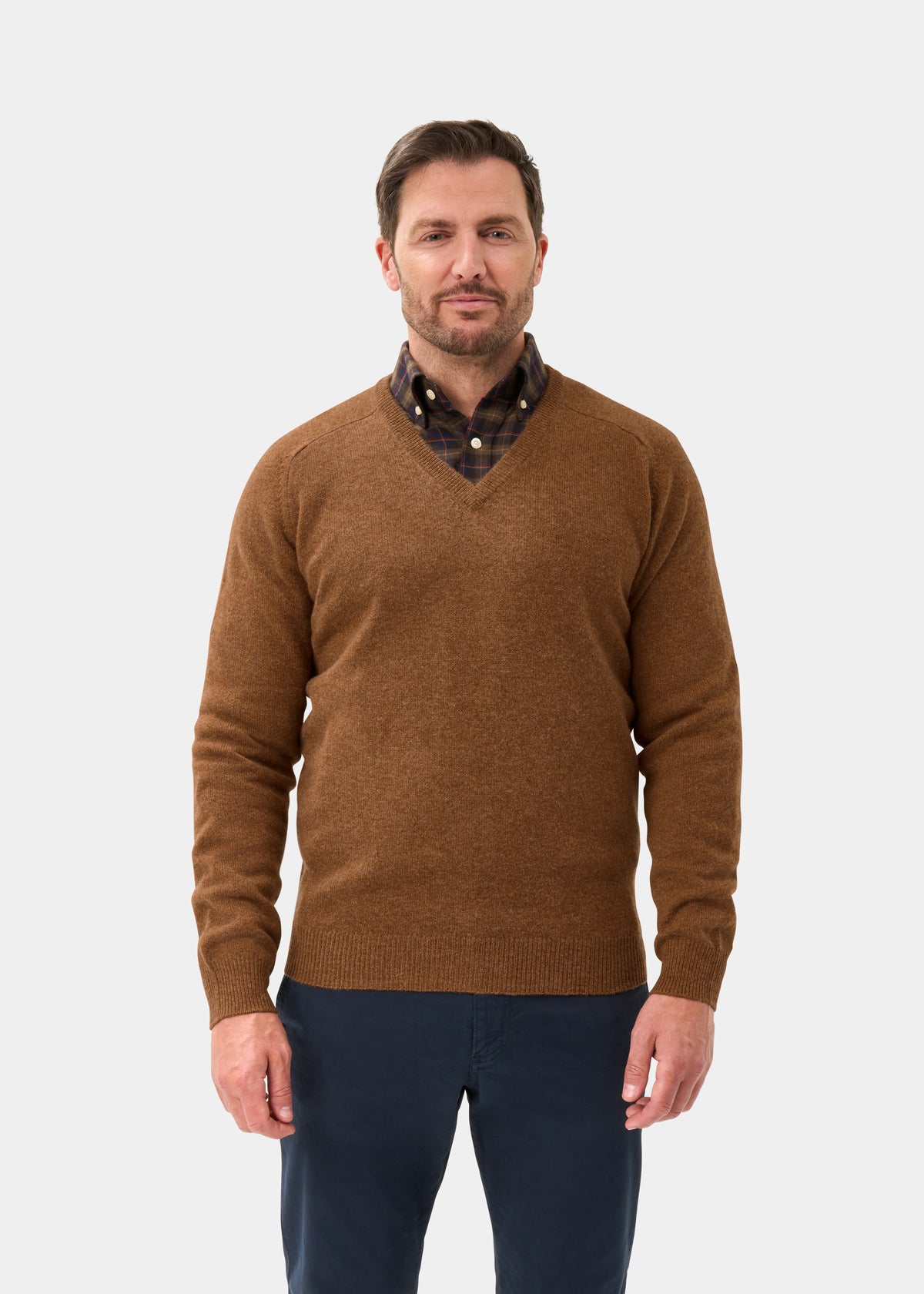 Man in Hampshire Lambswool Vee Neck Jumper in Driftwood, over plaid shirt and navy pants, on white background.