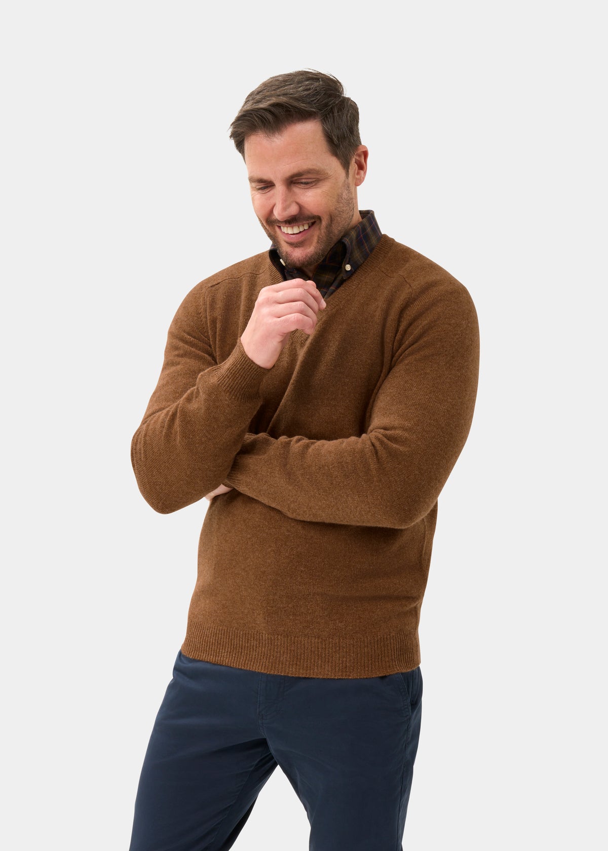 Man in Hampshire Lambswool Vee Neck Jumper In Driftwood smiles, looking down with one arm crossed near his mouth.